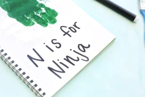Close-up of a spiral notebook on a pale blue surface, with green paint on the page and large black handwritten words visible.