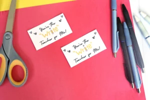 Desk scene with two white cards saying "You're the Write Teacher for Me!" surrounded by red background, hearts doodles, and writing supplies (scissors on left, pens on the right).