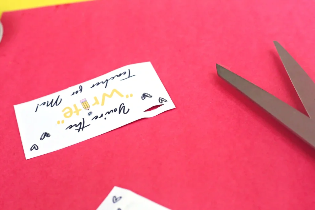 Scissors cutting a small white card with handwritten text and doodles on a bright pink surface