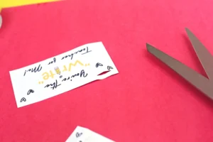 Scissors cutting a small white card with handwritten text and doodles on a bright pink surface