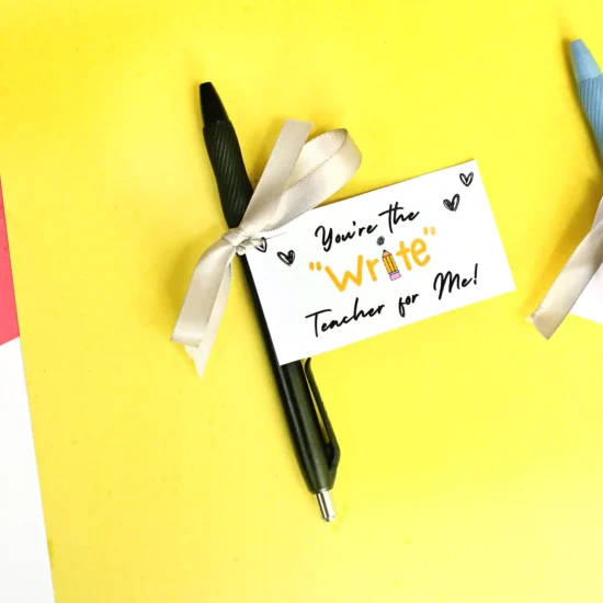 Pen with a white gift card reading "You're the Write teacher for Me!" on a bright yellow background, tied with a silver ribbon (teacher appreciation theme).