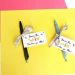 Two pens with ribbons attached to small gift cards that read 'You're the Write Teacher for Me!' on a yellow background with red paper in the corner.