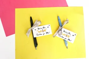 Two pens with ribbons attached to small gift cards that read 'You're the Write Teacher for Me!' on a yellow background with red paper in the corner.
