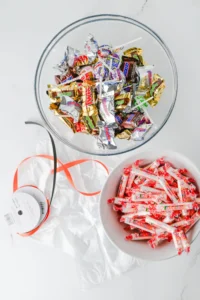 supplies for this classroom halloween treat- Top-down view of a glass bowl filled with assorted colorful wrapped candies on a white marble surface; some wrappers spill around the bowl and a second bowl with more candy is nearby.