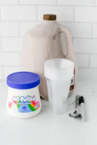 ingredients for the chocolate milk ghost cups: Jar of Jet-Puffed marshmallow creme on a kitchen counter, with a gallon of chocolate milk and clear cups