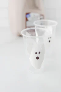 Close-up of a clear plastic cup filled with marshmallow fluff smears, with sharpie drawn eyes to look like ghosts for these halloween party drinks