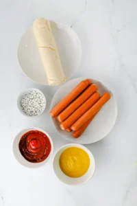 ingredients for this halloween dinner: Plate with several hot dogs on a white dish, crescent roll dough on a separate plate, and small bowls of condiments