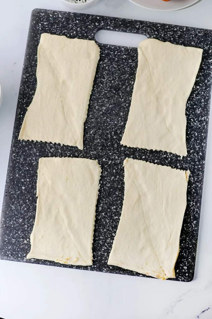 Mummy Hot Dogs - Cultivating Brilliant Minds Two elongated sheets of pale dough laid side by side on a dark speckled cutting board, ready to bake.