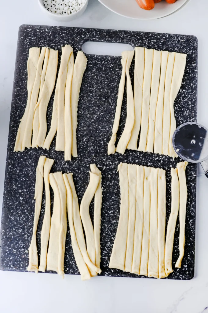 Mummy Hot Dogs - Cultivating Brilliant Minds Rows of flat dough ribbons laid in parallel on a dark speckled cutting board, with a round pastry cutter nearby.