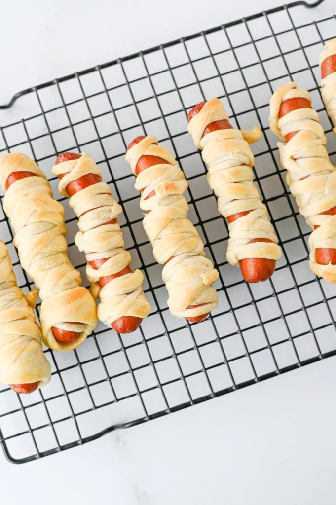 Mummy Hot Dogs - Cultivating Brilliant Minds hot dogs wrapped in pale pastry laid on a metal cooling rack for a halloween lunch