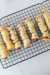 Mummy-style hot dogs wrapped in dough with googly eyes on a cooling rack.