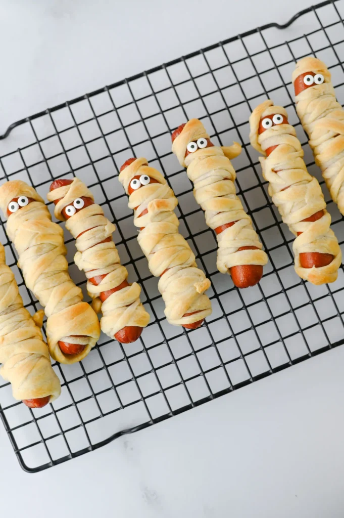 Mummy Hot Dogs - Cultivating Brilliant Minds Mummy-style hot dogs wrapped in dough with googly eyes on a cooling rack.