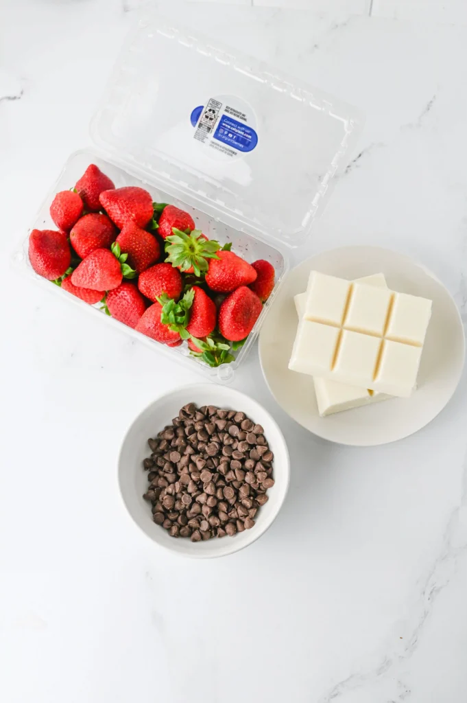 ingredients for the chocolate covered strawberry ghosts: Fresh strawberries in a clear container beside a plate of white chocolate squares and a bowl of chocolate chips on a marble surface.