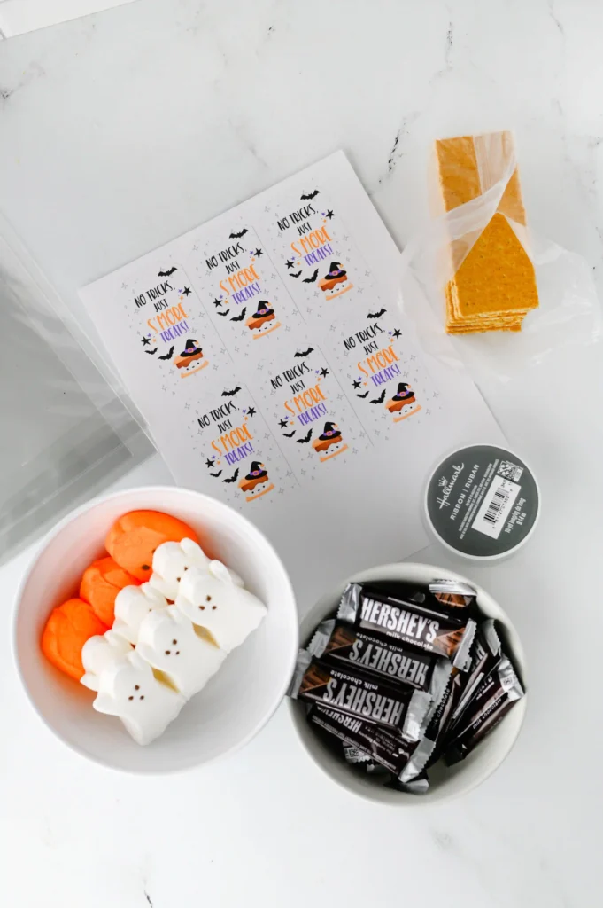 Halloween S'mores Treat Bags - Cultivating Brilliant Minds supplies for the halloween s'mores treat bags: Assorted Halloween treats on a marble surface: printed cards with spooky designs, marshmallow treats in orange and white, and wrapped Hershey's chocolates nearby.
