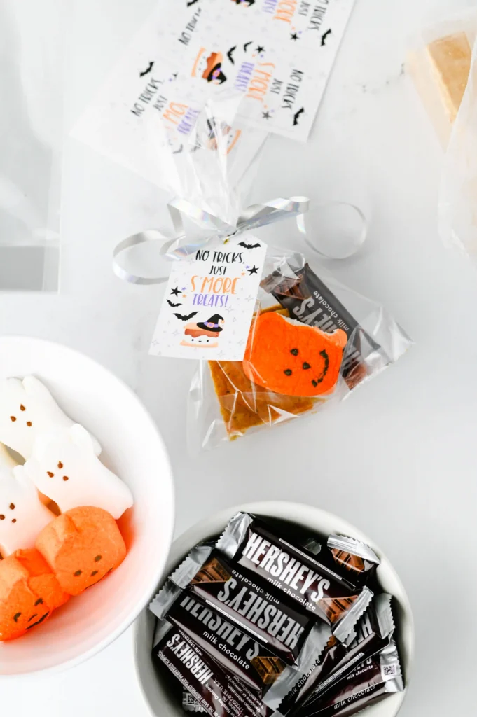 Halloween S'mores Treat Bags - Cultivating Brilliant Minds Halloween-themed treat assortment: pumpkin-faced peeps, Hershey's bars, graham crackers and gift tags reading 'No tricks, just S’more treats'.