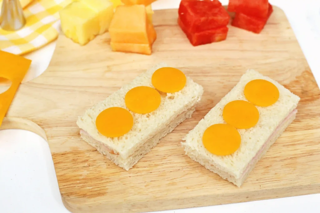 LEGO Lunchbox Idea - Cultivating Brilliant Minds Sliced sandwiches on a wooden board topped with bright orange cheese pieces; in the background are cheese cubes and a bottle cap on a checkered cloth.