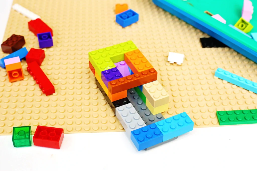 Scattered colorful LEGO bricks on a beige studded baseplate with a partially built multicolor structure nearby and a blue-green piece in the background.