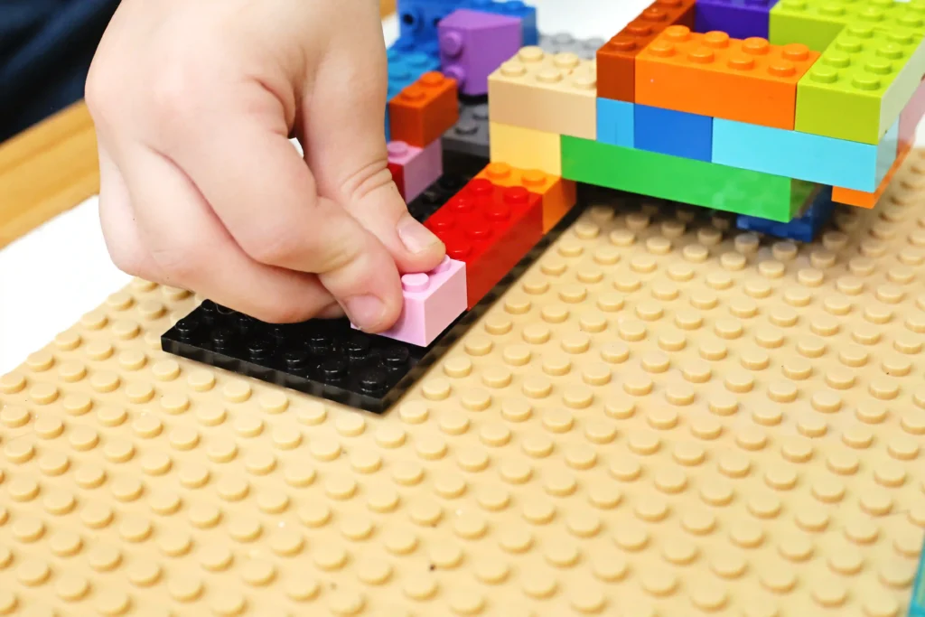 Hand placing a light pink LEGO brick onto a black base plate as a colorful LEGO structure sits nearby.