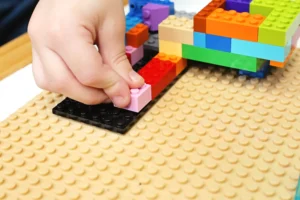 Hand placing a light pink LEGO brick onto a black base plate as a colorful LEGO structure sits nearby.