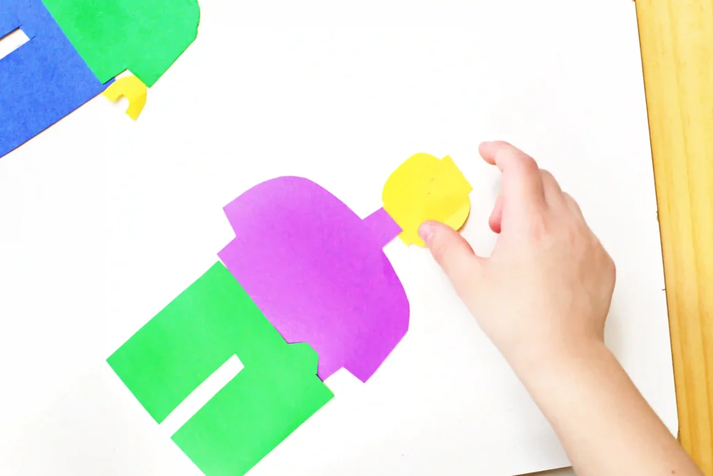 LEGO Minifigures - Cultivating Brilliant Minds Overhead view of a craft project with green, blue, purple, and yellow paper shapes on a white surface; a hand places a yellow piece.