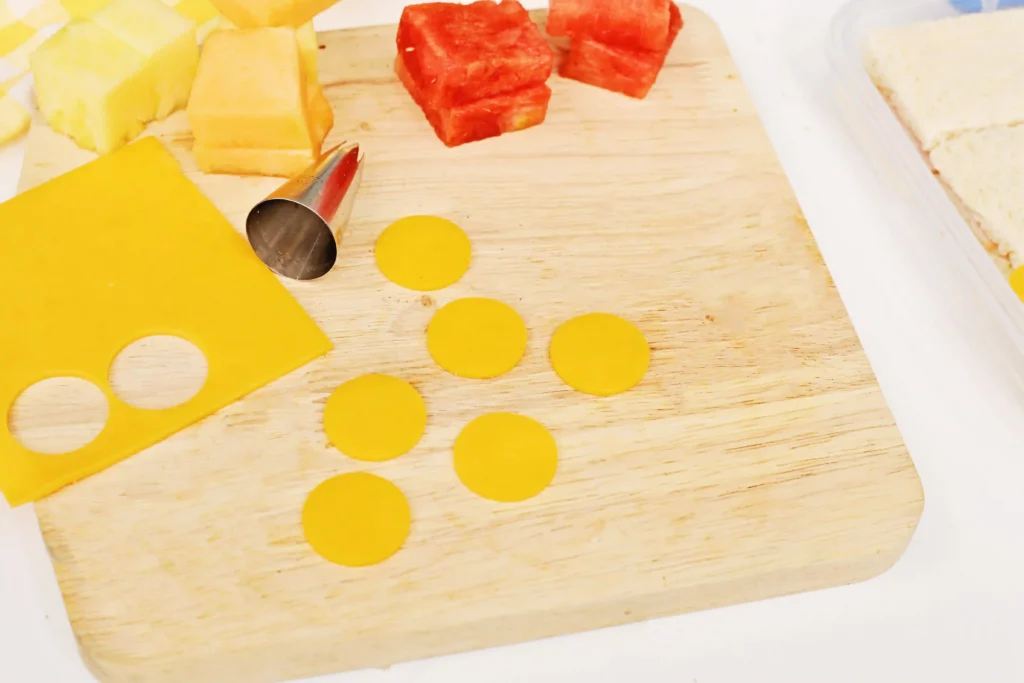 LEGO Lunchbox Idea - Cultivating Brilliant Minds Cheese on a wooden cutting board: yellow cheddar cubes and rounds, red cheese cubes, a metal round cookie cutter, and scattered circular cheese slices