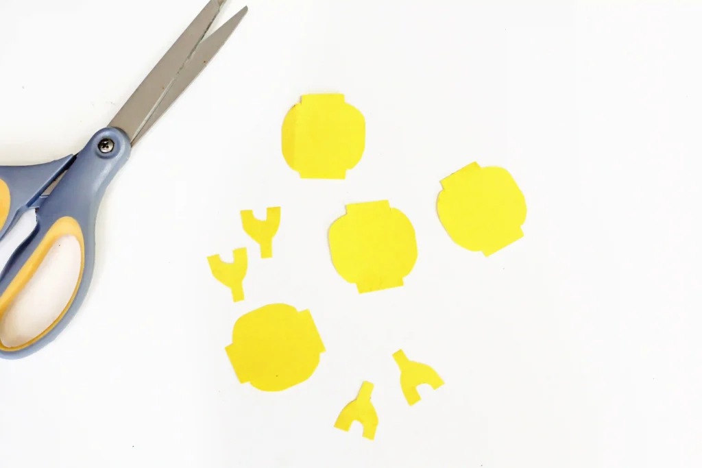 LEGO Minifigures - Cultivating Brilliant Minds Yellow paper cutouts in abstract rounded shapes scattered on a white surface, with craft scissors at the left.