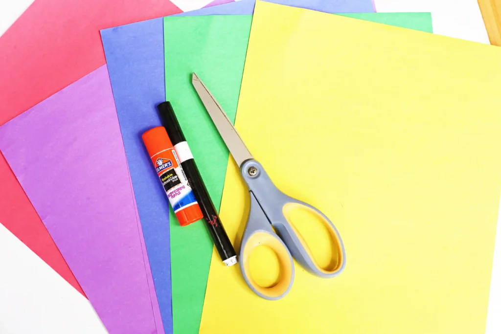 LEGO Minifigures - Cultivating Brilliant Minds supplies for the LEGO minifigure craft: Bright assortment of construction paper in pink, purple, blue, green, and yellow with scissors and a glue stick laid on top, ready for a craft project.