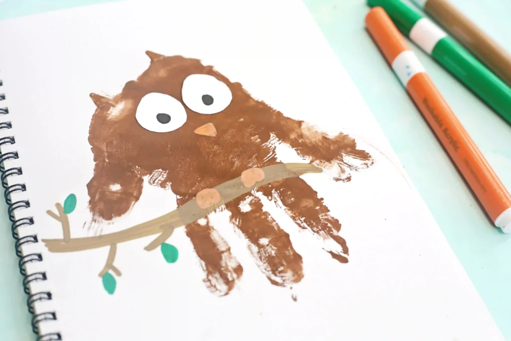 Brown handprint forms a cartoon owl face with large white eyes and a small orange nose/beak on a spiral notebook, with markers nearby.