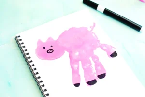 Pink pig handprint painting on a spiral-bound notebook page with a black marker nearby.