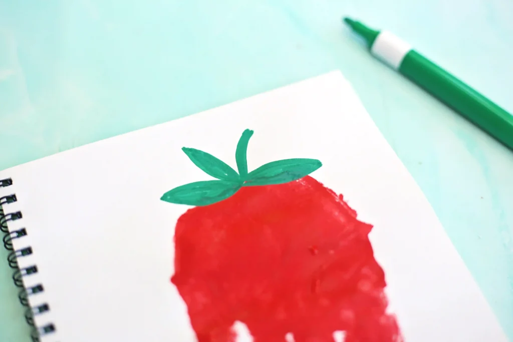 Close-up of a spiral notebook page with a red-painted strawberry and green leaves, a green marker on a mint background to the right.