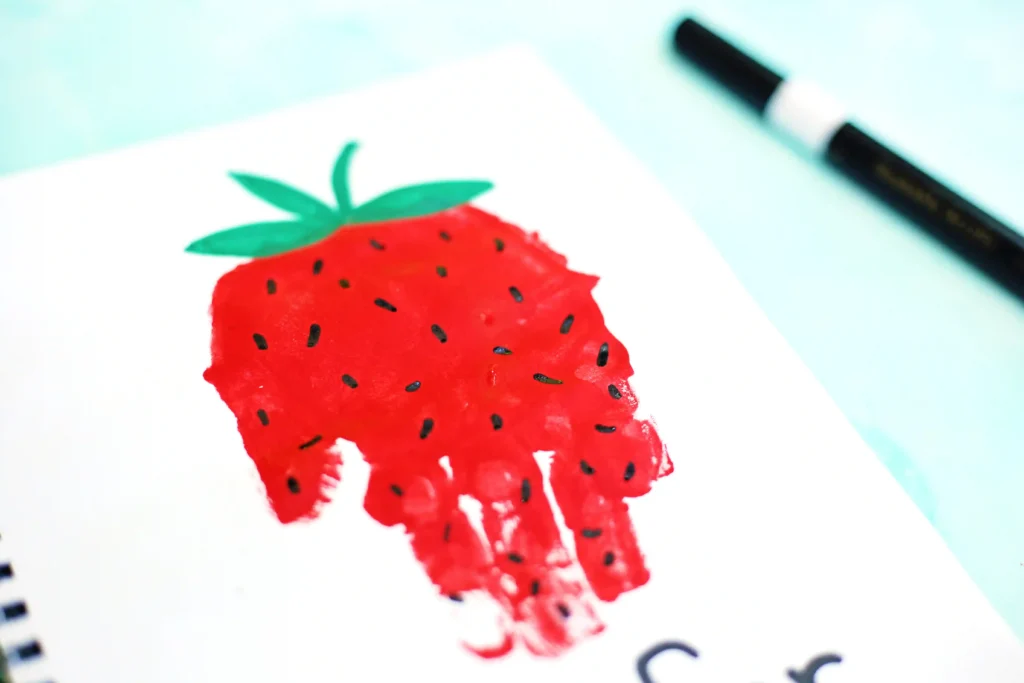 Close-up of a bright red strawberry drawing on white paper with green leaves, a black marker in the background.