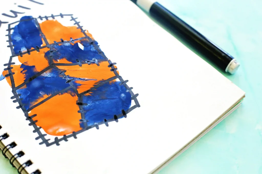Close-up of a notebook page with orange and blue handprint within a stitched grid pattern and a pen nearby.