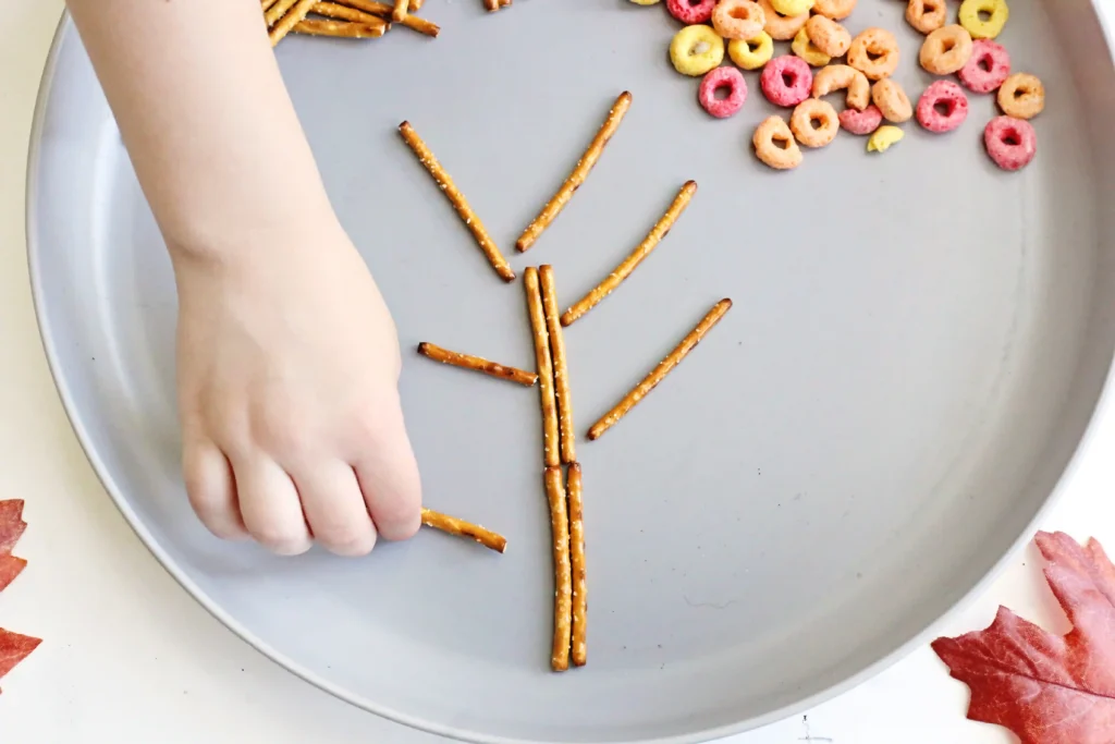 Child's hand places pretzel sticks on a round gray tray, with colorful ring-shaped cereal clustered in the corner, creating snack art.