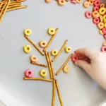 Plate with pretzel sticks arranged to form a stick-figure person, surrounded by colorful ring-shaped cereals; a hand is placing a cereal ring, as if completing the artwork.