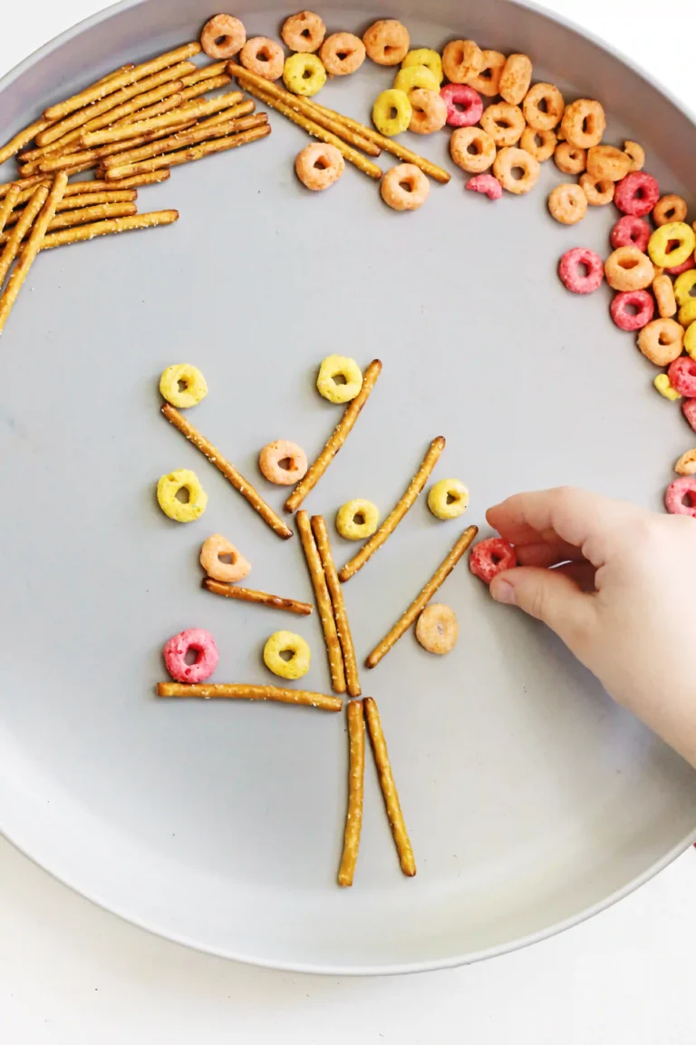 Plate with pretzel sticks arranged to form a stick-figure person, surrounded by colorful ring-shaped cereals; a hand is placing a cereal ring, as if completing the artwork.