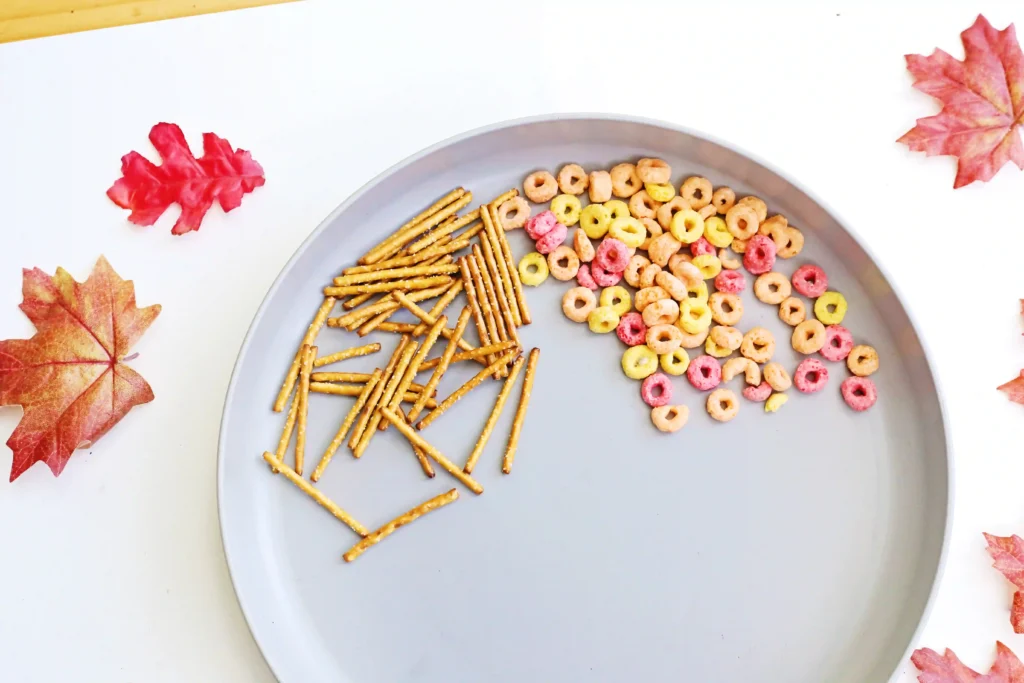 supplies for this edible fall tree craft: Gray plate holds a snack mix of colorful ring cereal and pretzel sticks, with autumn leaves around the edge of the image.