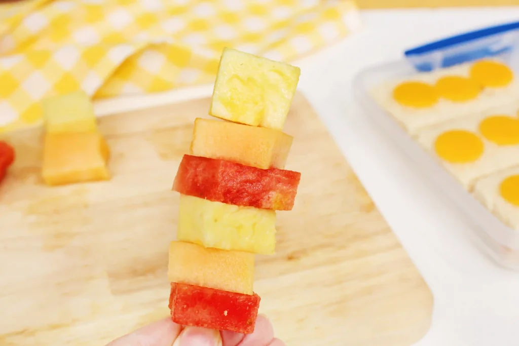 LEGO Lunchbox Idea - Cultivating Brilliant Minds Person holds a skewer of alternating watermelon and cantaloupe cubes, close-up on the fruit stack.