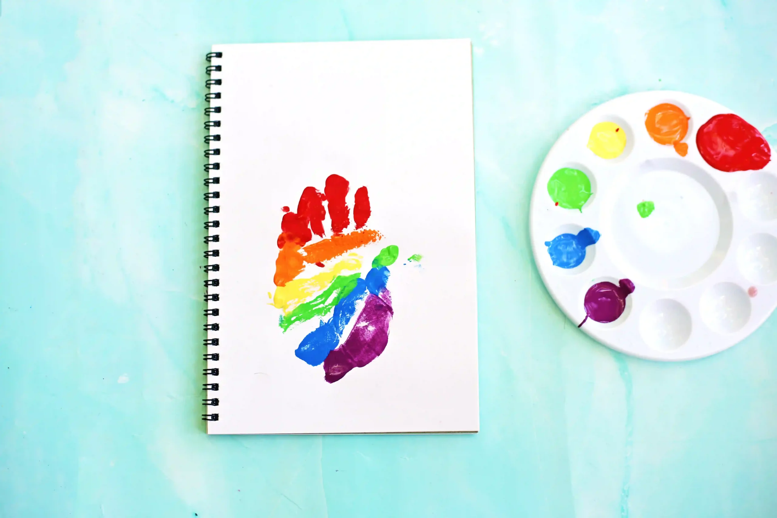 Spiral notebook with a rainbow handprint on the page, resting beside a painter's color palette on a light turquoise surface.