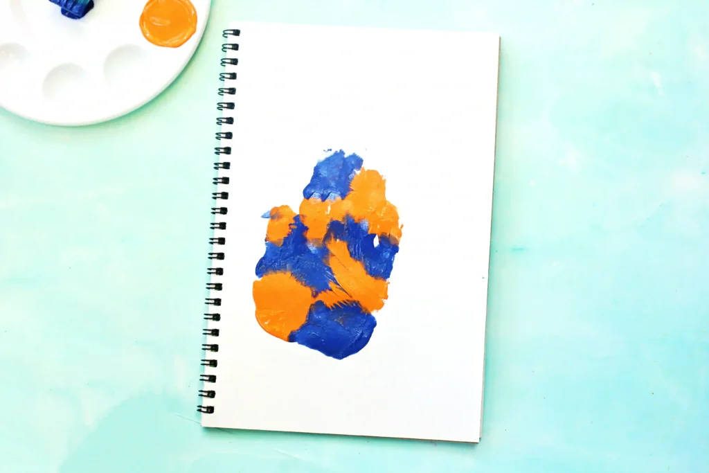 Spiral notebook opened on a mint surface with orange and blue paint splotches on the page; a painter's palette sits in the corner above left.