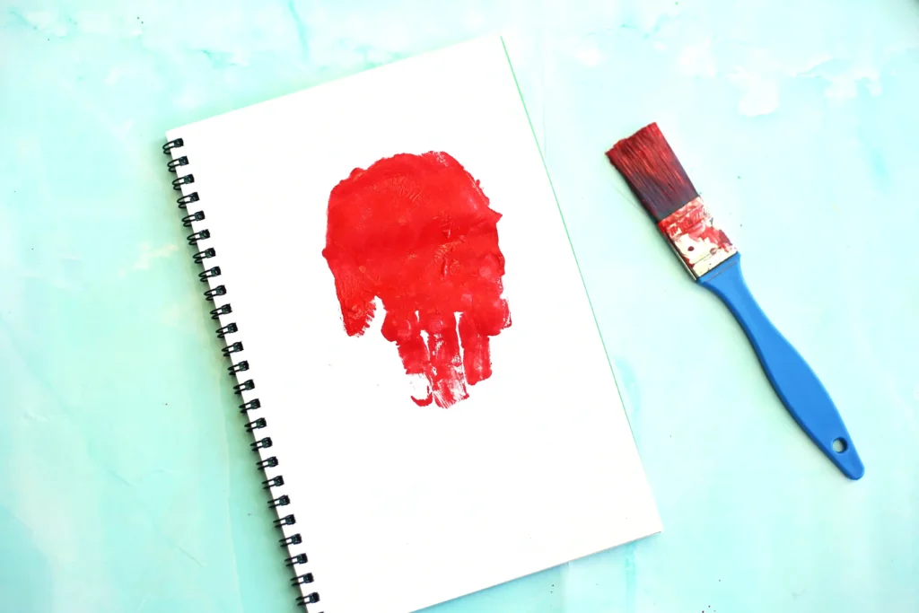 Red handprint painted on a white notebook page with a blue-handled paintbrush nearby on a mint background.