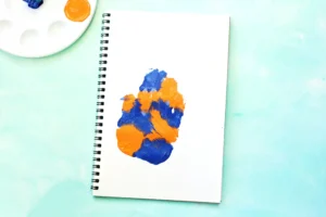 Spiral notebook opened on a mint surface with orange and blue paint splotches on the page; a painter's palette sits in the corner above left.