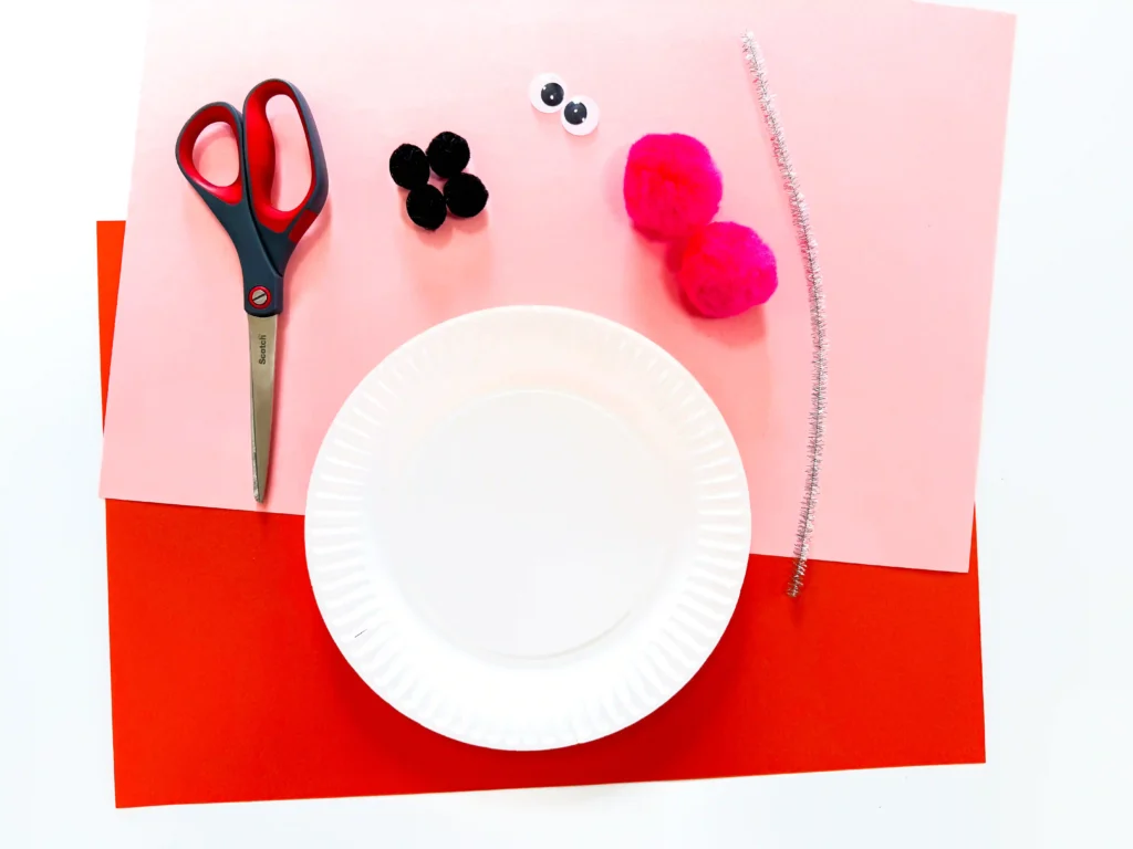 Paper Plate Snowman Craft - Cultivating Brilliant Minds supplies for the paper plate snowman craft: pink and orange paper, scissors, black pom-poms, googly eyes, two pink pom-poms, a white paper plate, and a silver pipe cleaner