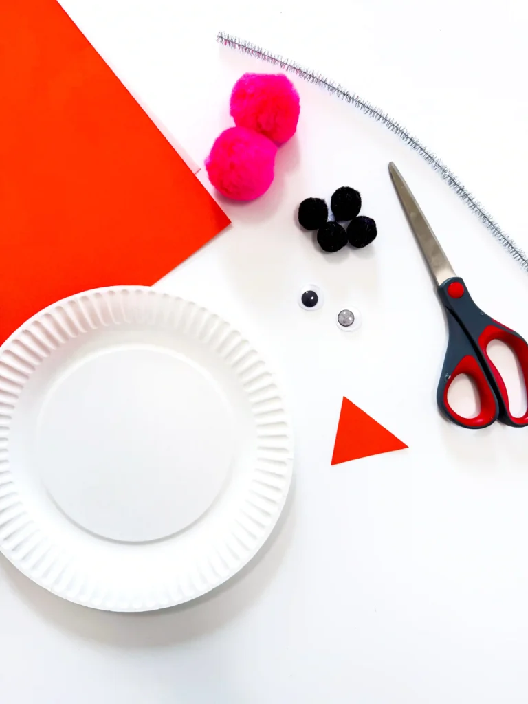 Paper Plate Snowman Craft - Cultivating Brilliant Minds Craft supplies on a white surface: red cardstock, pink and black pom poms, scissors, a white paper plate, and red paper shapes.
