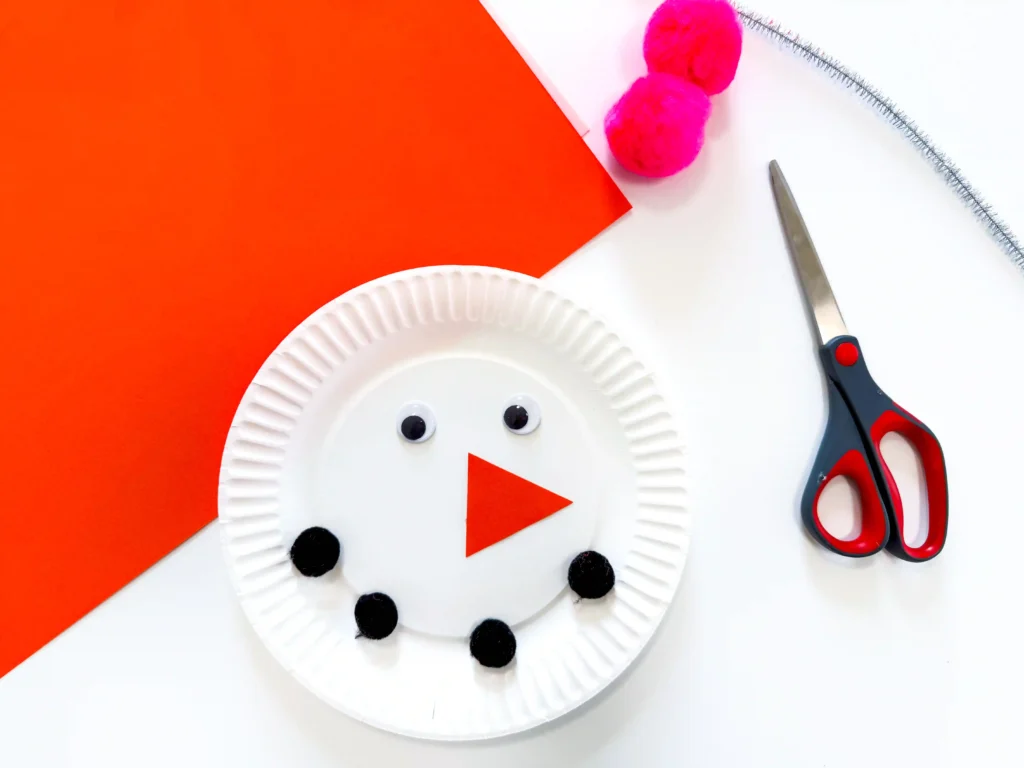 Paper Plate Snowman Craft - Cultivating Brilliant Minds DIY snowman craft using a paper plate with googly eyes and black pom-pom buttons on an orange background; scissors and pink pom-poms nearby.