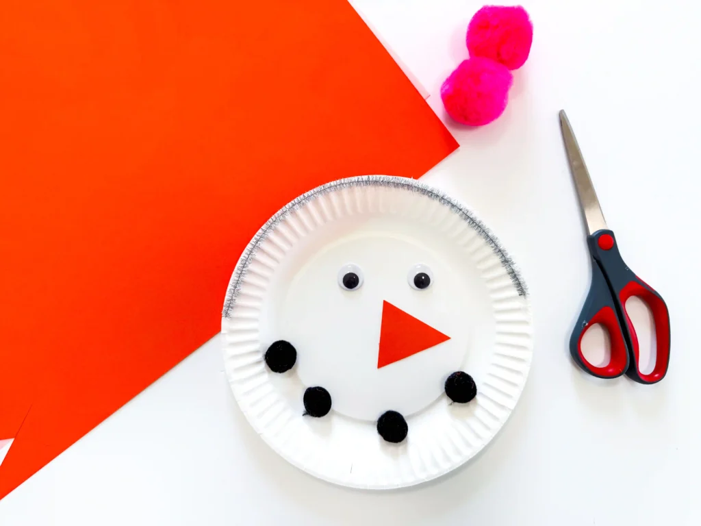 Paper Plate Snowman Craft - Cultivating Brilliant Minds Crafty winter scene: white paper plate snowman with black buttons and silve pipe cleaner, pink pom-poms, and scissors on an orange and white background.