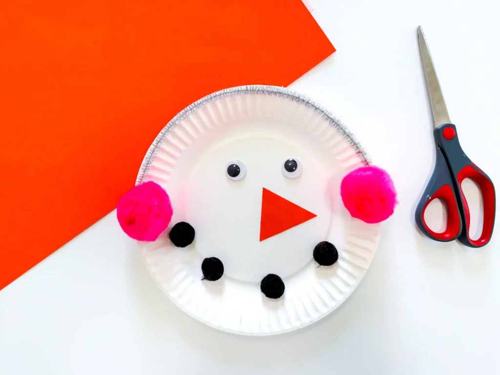 Paper Plate Snowman Craft - Cultivating Brilliant Minds Paper-plate snowman craft with googly eyes, pink earmuffs, black buttons, and an orange triangle nose on an orange background, with scissors nearby.
