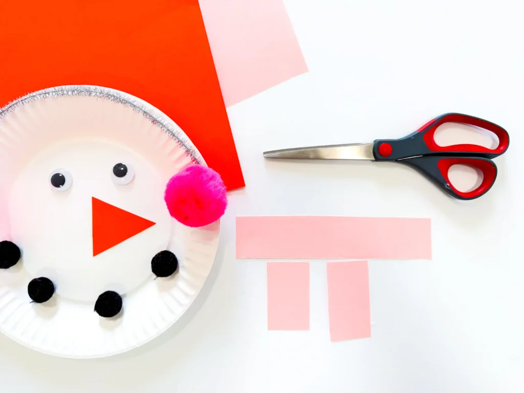 Paper Plate Snowman Craft - Cultivating Brilliant Minds Craft project scene: scissors on a white surface beside pink construction paper cut into strips
