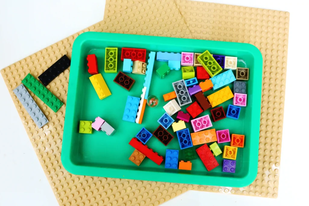 Colorful LEGO bricks scattered in a green tray on a tan studded baseplate backdrop