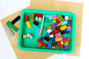 Colorful LEGO bricks scattered in a green tray on a tan studded baseplate backdrop; a small building block assortment inside the tray prize.