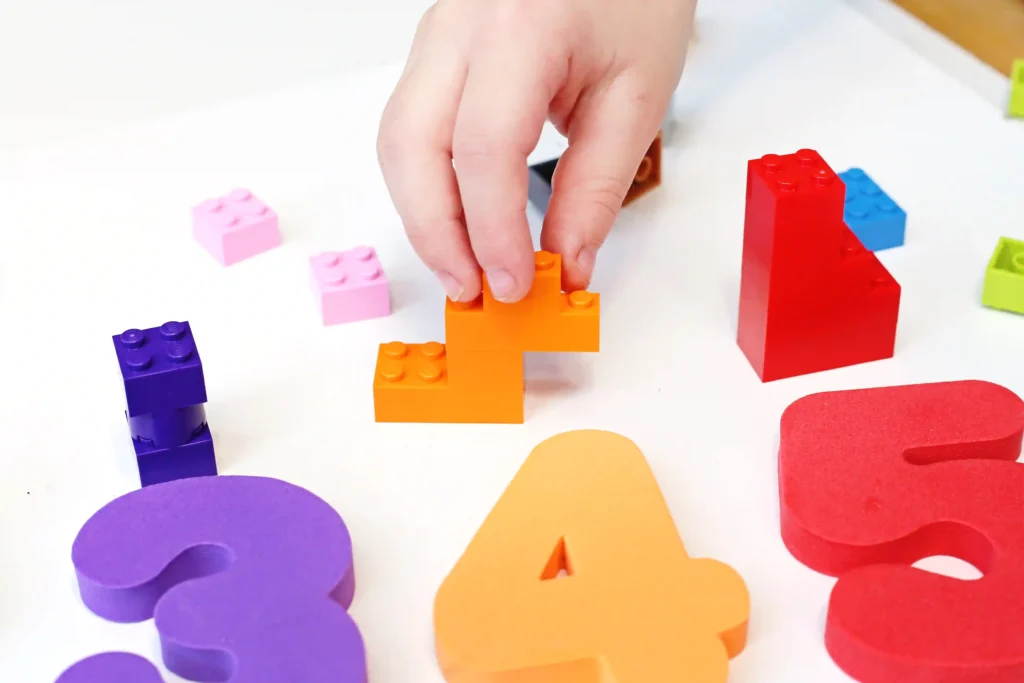 Child's hand stacks orange LEGO bricks among scattered purple, pink, and red pieces on a white surface.
